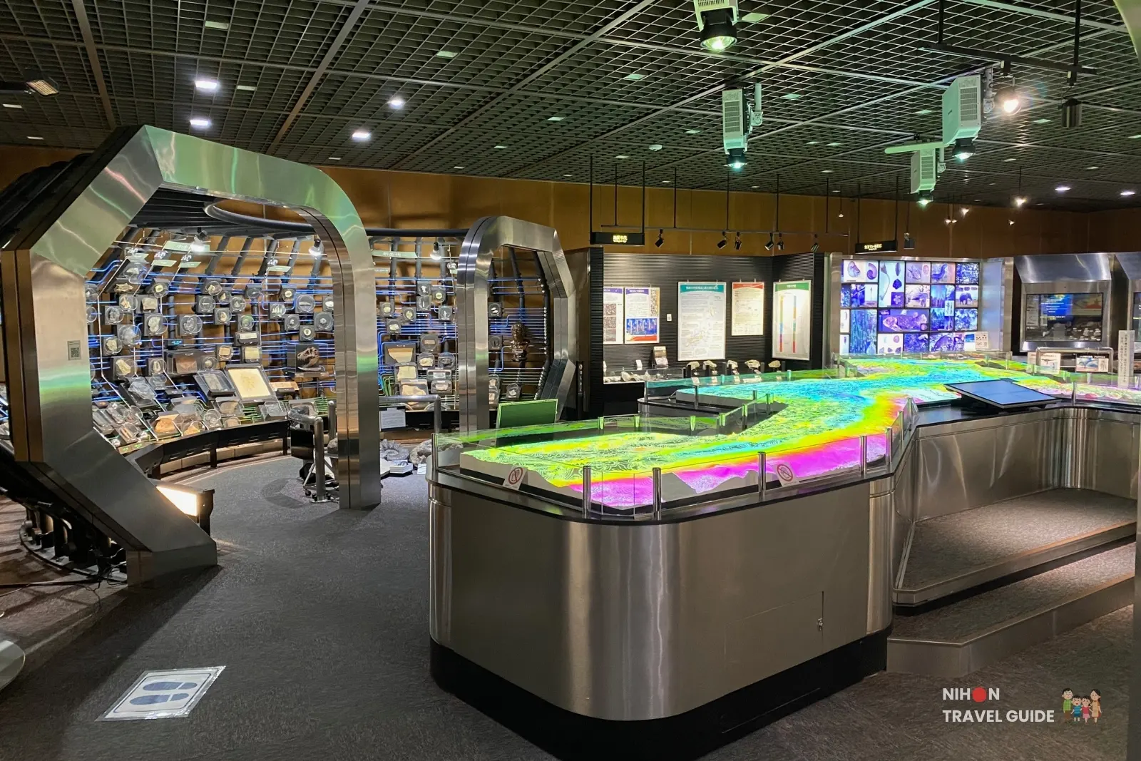 Exhibition Room 1 at the Tsukuba Geological Museum featuring the large illuminated 3D geological model of the Japanese Islands with colorful projection mapping, surrounded by specimen displays and interactive panels.