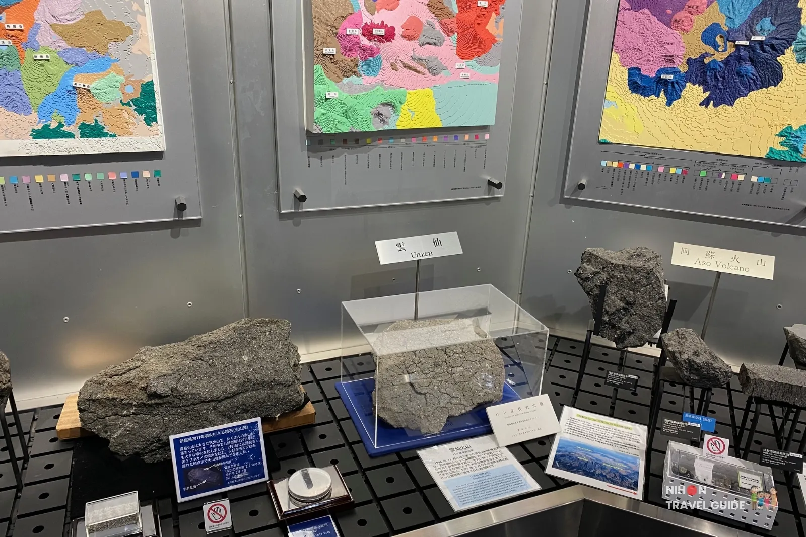 Close-up of rock specimens from Unzen and Aso volcanoes displayed in Exhibition Room 3 of the Tsukuba Geological Museum, with colorful geological maps in the background.