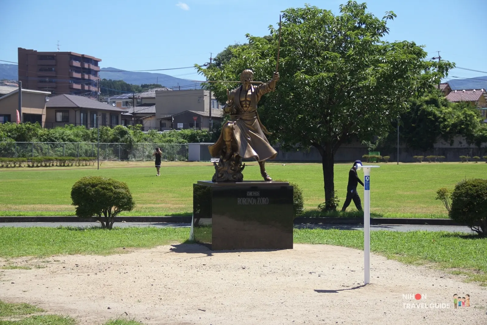 Side-front view of the bronze Roronoa Zoro statue from One Piece standing in Ozu Central Park under a clear blue sky in Kumamoto.