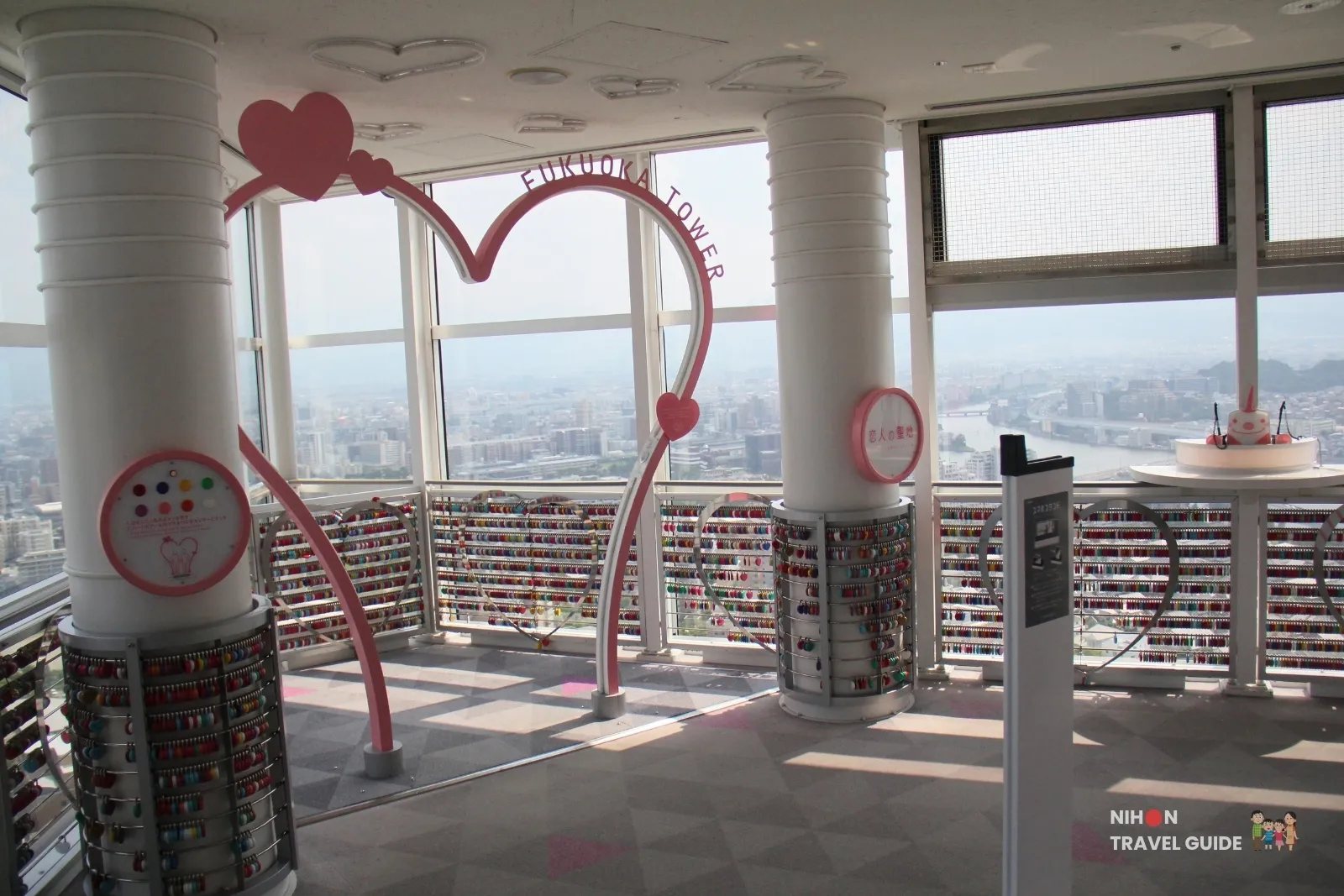 Pink Fukuoka Tower heart arch at Lover’s Sanctuary on observation 1F, with love locks on the railings and Fukuoka city in the background.