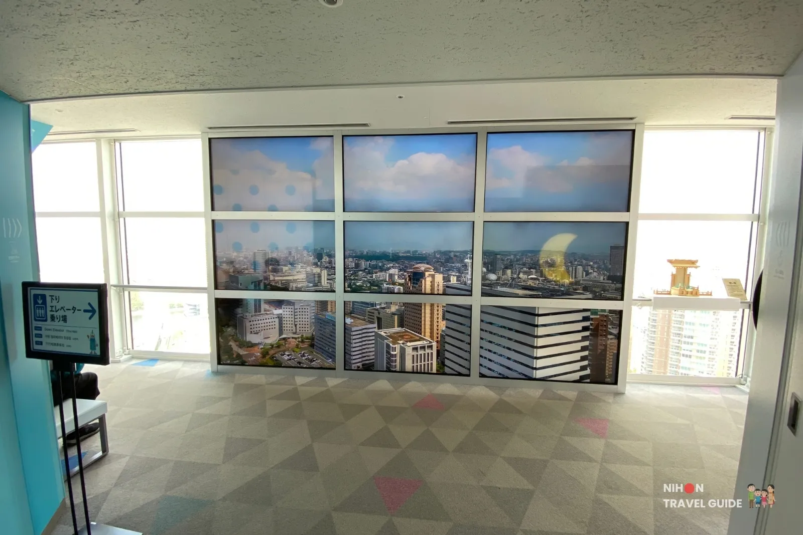 Nine-screen Magic Window video wall on the Fukuoka Tower observation deck showing a live-style city panorama next to the down-elevator sign.
