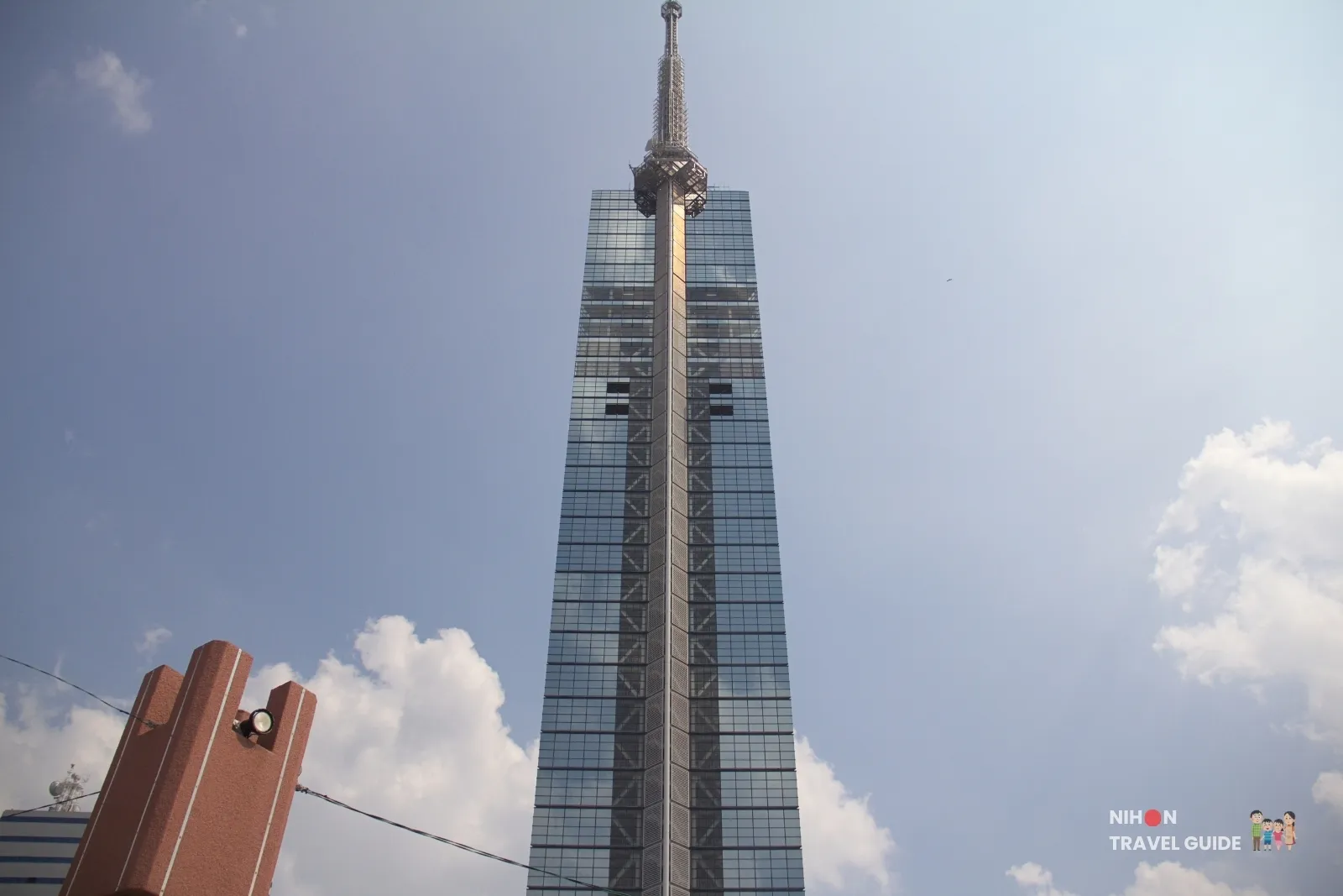 Low-angle daytime view of Fukuoka Tower’s triangular glass Mirror Sail facade and antenna against a blue sky with clouds, photographed from street level in Momochihama.