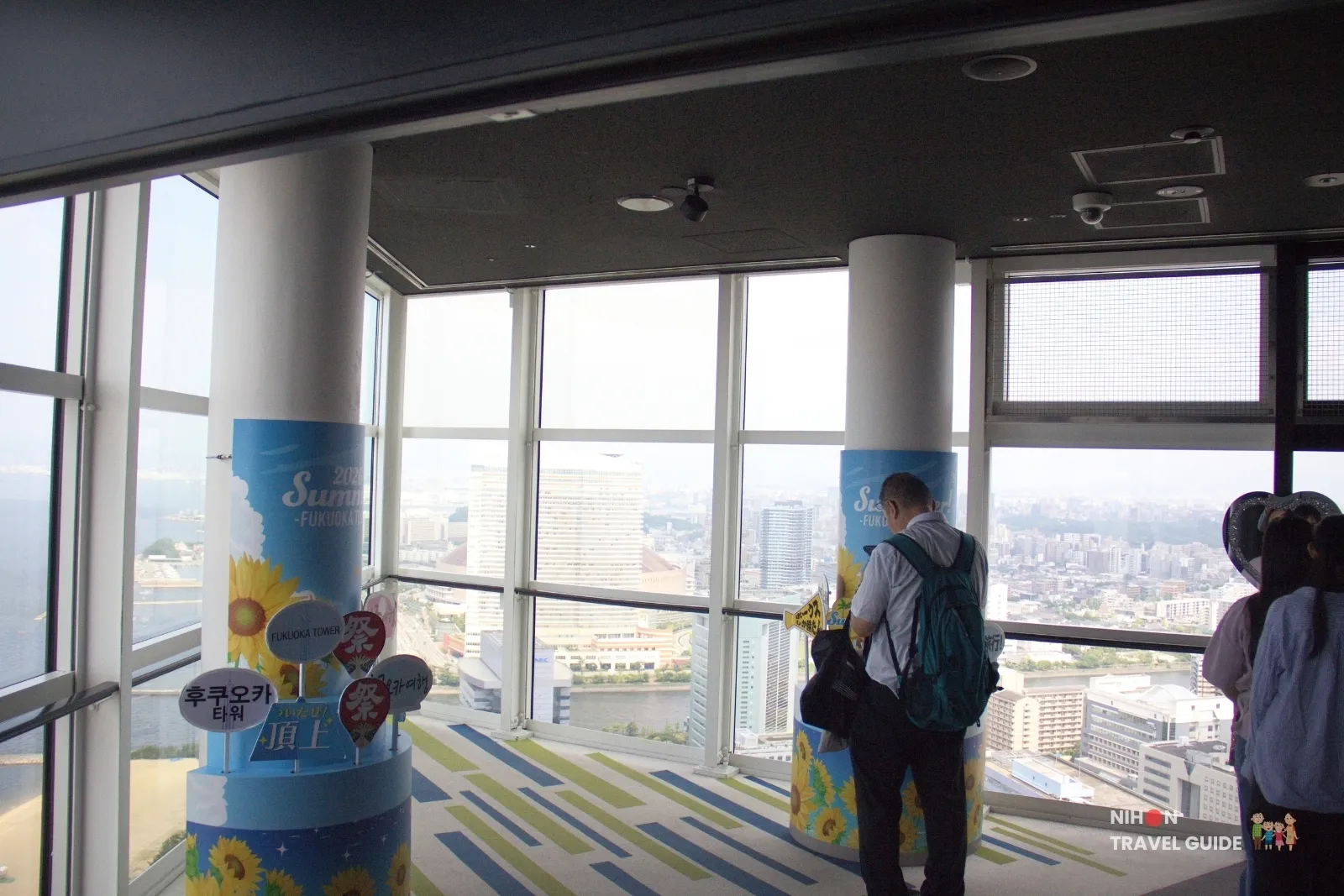 Photo-prop corner on the Fukuoka Tower observation deck with 2026 Summer sunflower wraps and visitors looking over the city.