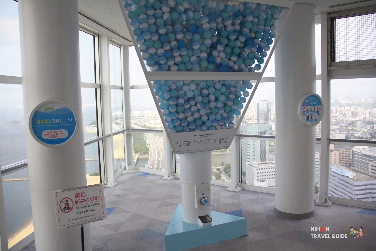 Giant Sky Gacha capsule machine filled with blue and white balls on Fukuoka Tower observation 1F, with Hakata Bay visible through the windows.