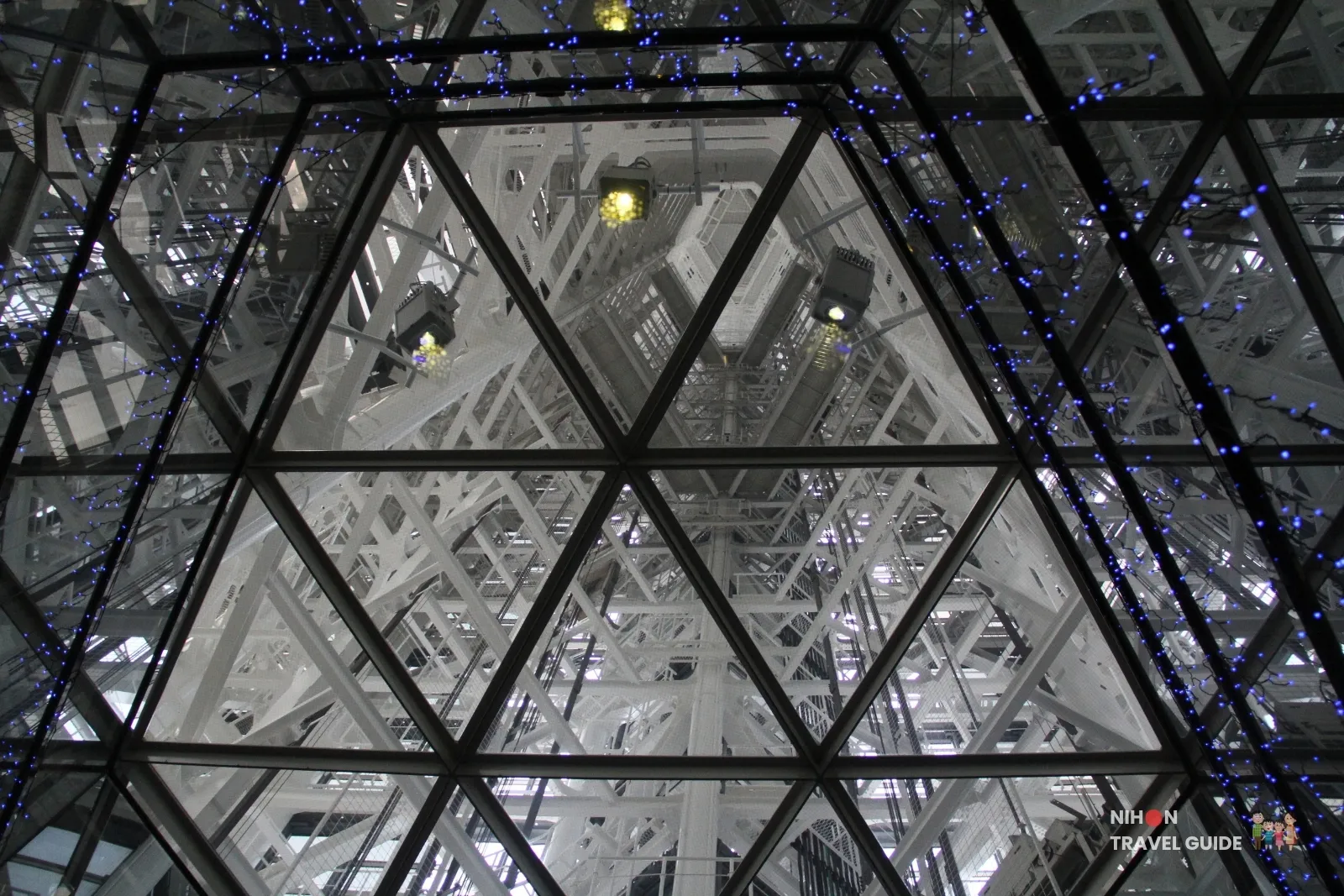 Looking up through the glass ceiling of Fukuoka Tower’s SKY Port elevator lobby at the white steel lattice and blue LED lights inside the shaft.