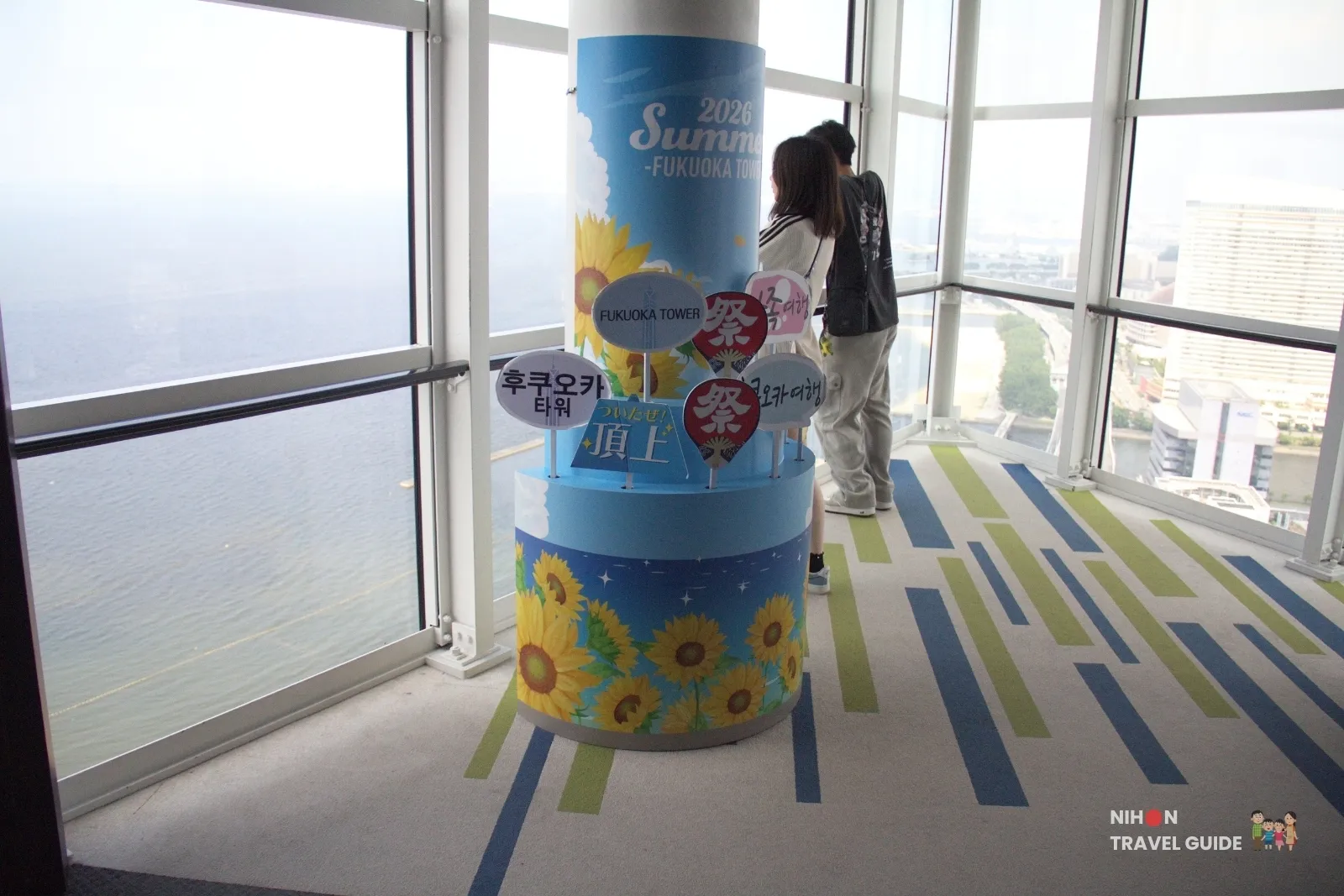 Summer 2026 sunflower photo props and handheld signs on a Fukuoka Tower observation-deck column beside Hakata Bay windows.