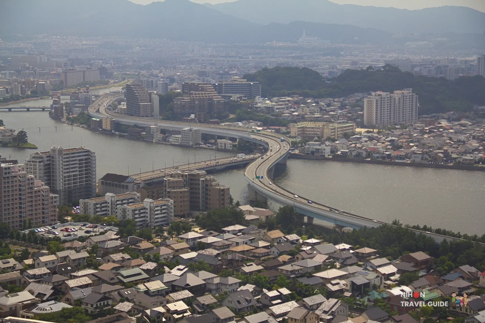 View from Fukuoka Tower of the Fukuoka Urban Expressway curving over the Muromi River, with houses, a wooded hill, and mountains behind.