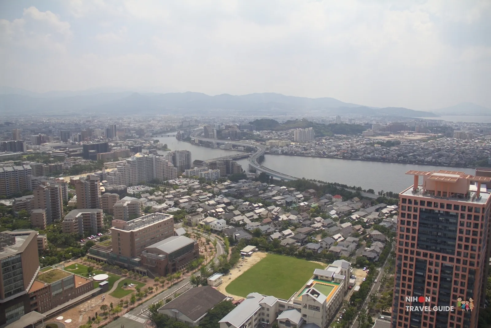 View from Fukuoka Tower south inland over Fukuoka City Library and a sports field, a river, the urban expressway, and mountains beyond the suburbs.