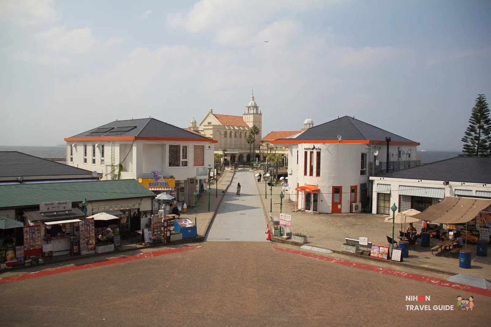 Walkway through Momochi Seaside Park toward Marizon on Hakata Bay, with Blue Seal ice cream and white seaside shops lining the path.