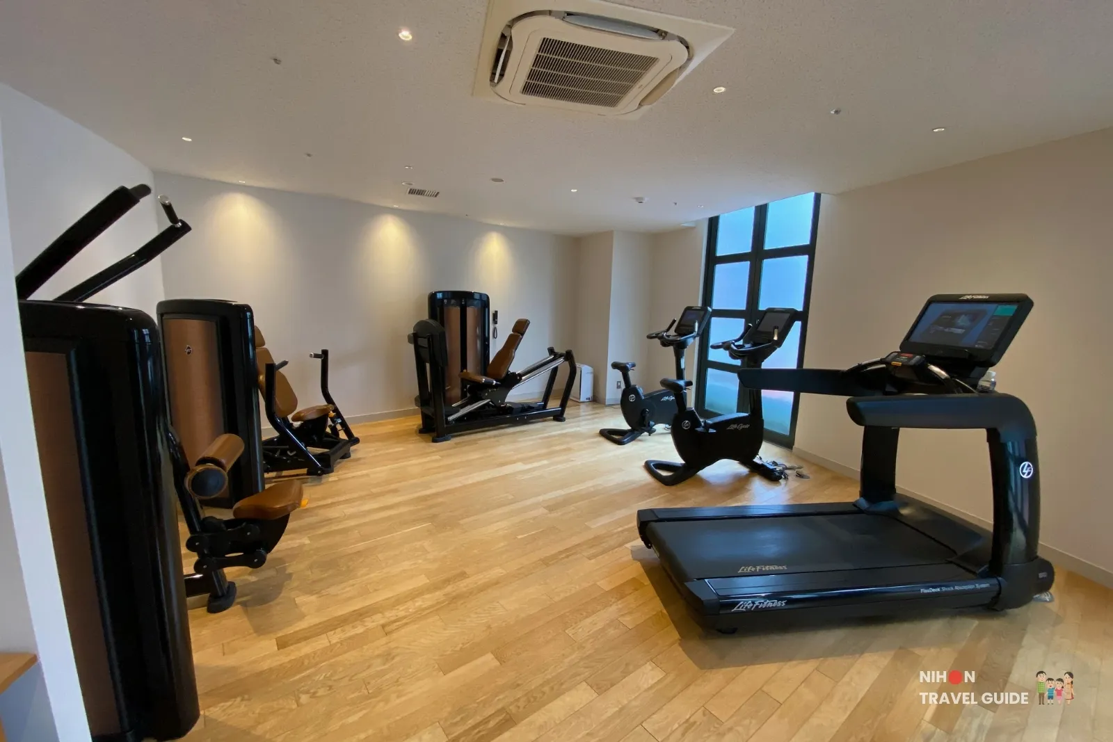 Guest fitness room at THE BASICS FUKUOKA with Life Fitness treadmill, exercise bikes, and strength machines on a wood floor beside a tall window.