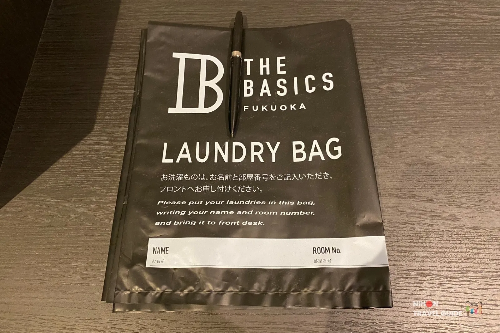Black THE BASICS FUKUOKA laundry bag on a desk with a pen, bilingual drop-off instructions, and blank fields for guest name and room number.