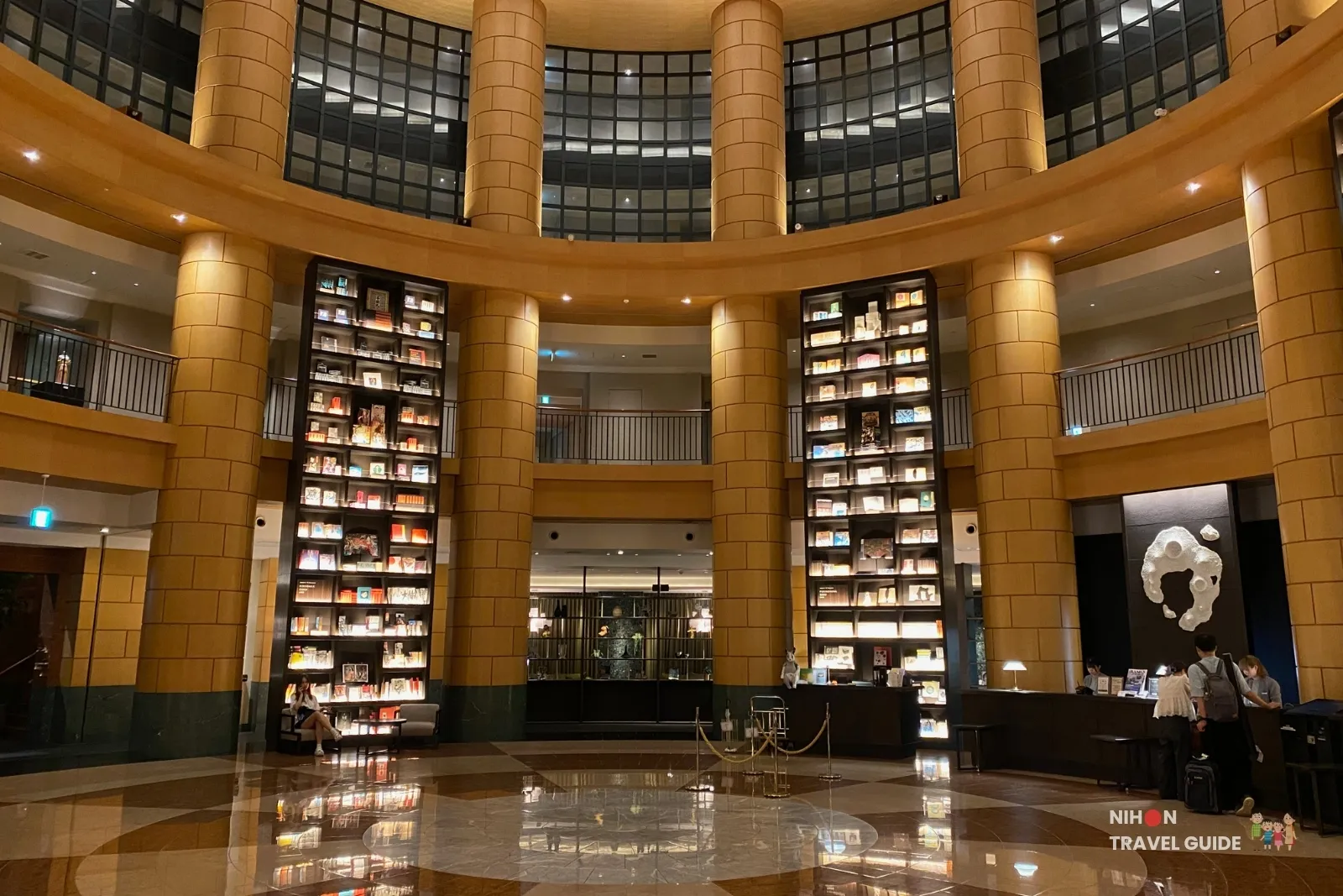 Circular atrium lobby at THE BASICS FUKUOKA with tall illuminated bookshelves between stone columns, marble floor, and reception desk on the right.