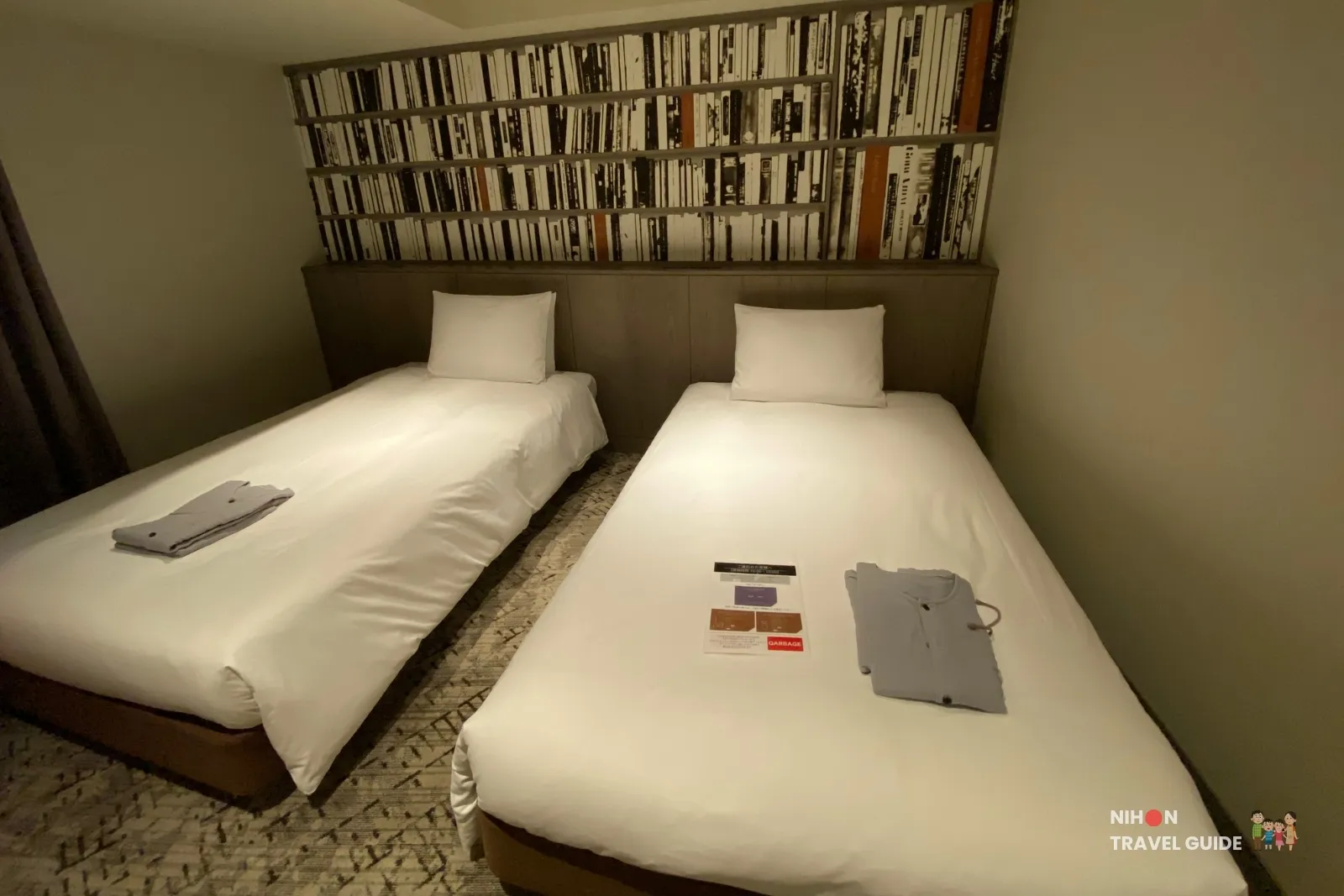 Two single beds in a THE BASICS FUKUOKA Moderate Twin with white linens, folded gray nightwear, a housekeeping card, and a bookshelf-print headboard.