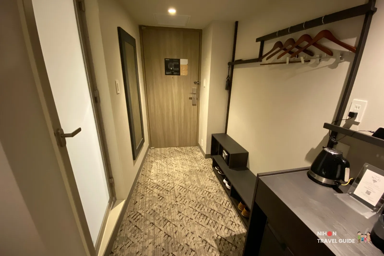 Entry hallway of a THE BASICS FUKUOKA Moderate Twin showing the guest door, full-length mirror, open hanging rail with wooden hangers, shoe shelf, and kettle console.