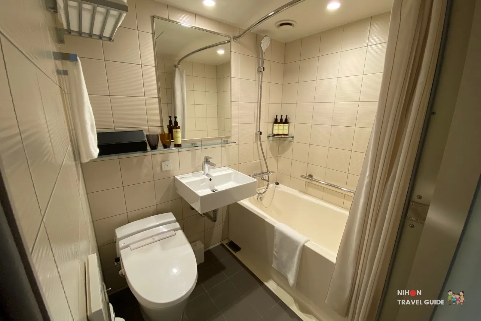 Compact unit bathroom at THE BASICS FUKUOKA with washlet toilet, wall-mounted sink, mirror shelf, and bathtub-shower behind a curtain.