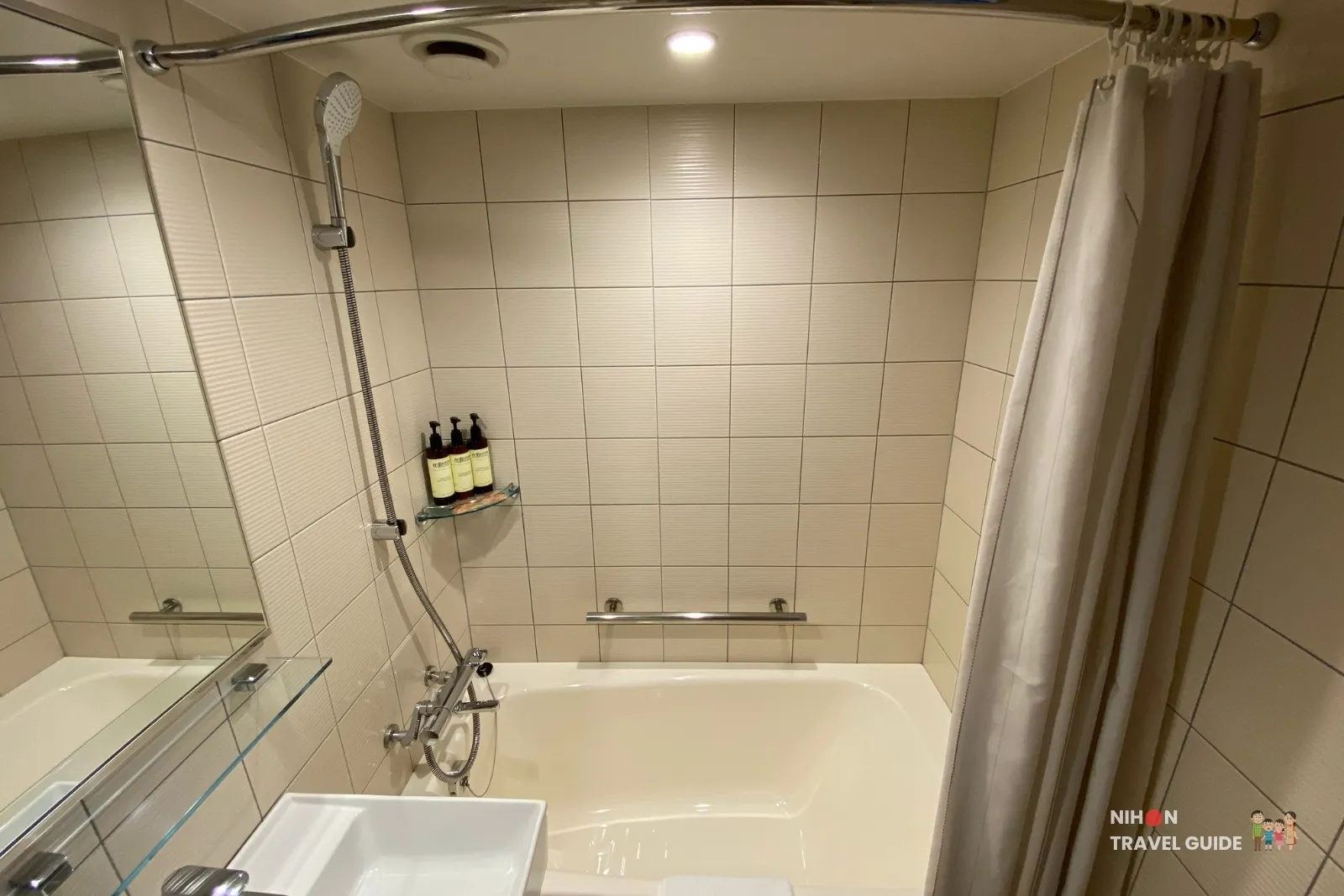 Beige-tiled unit bathroom at THE BASICS FUKUOKA with a deep Japanese bathtub, handheld shower, grab bar, and pump bottles on a glass corner shelf.