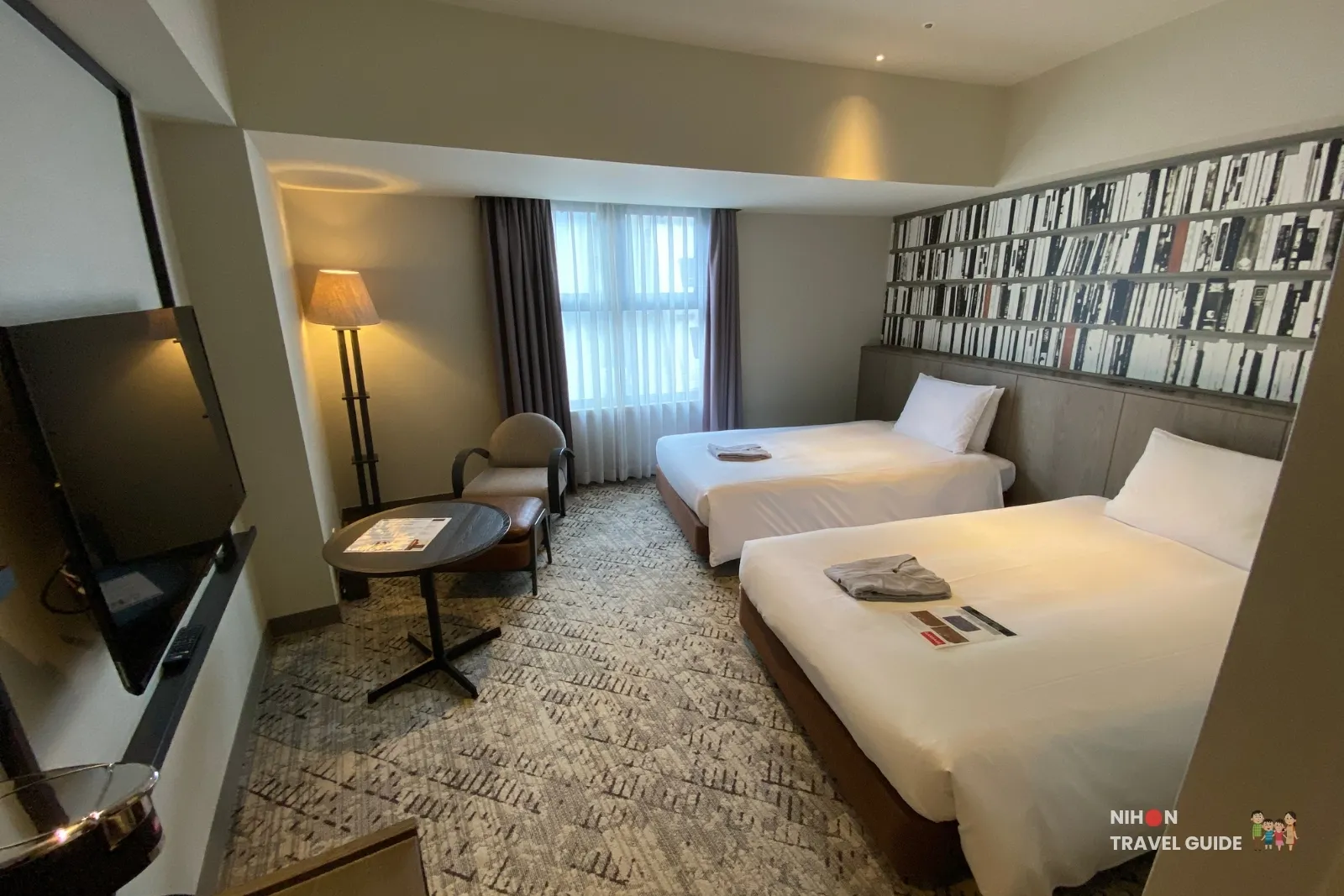 Overview of a THE BASICS FUKUOKA Moderate Twin with two beds, armchair and ottoman by the window, floor lamp, round side table, wall-mounted TV, and patterned carpet.
