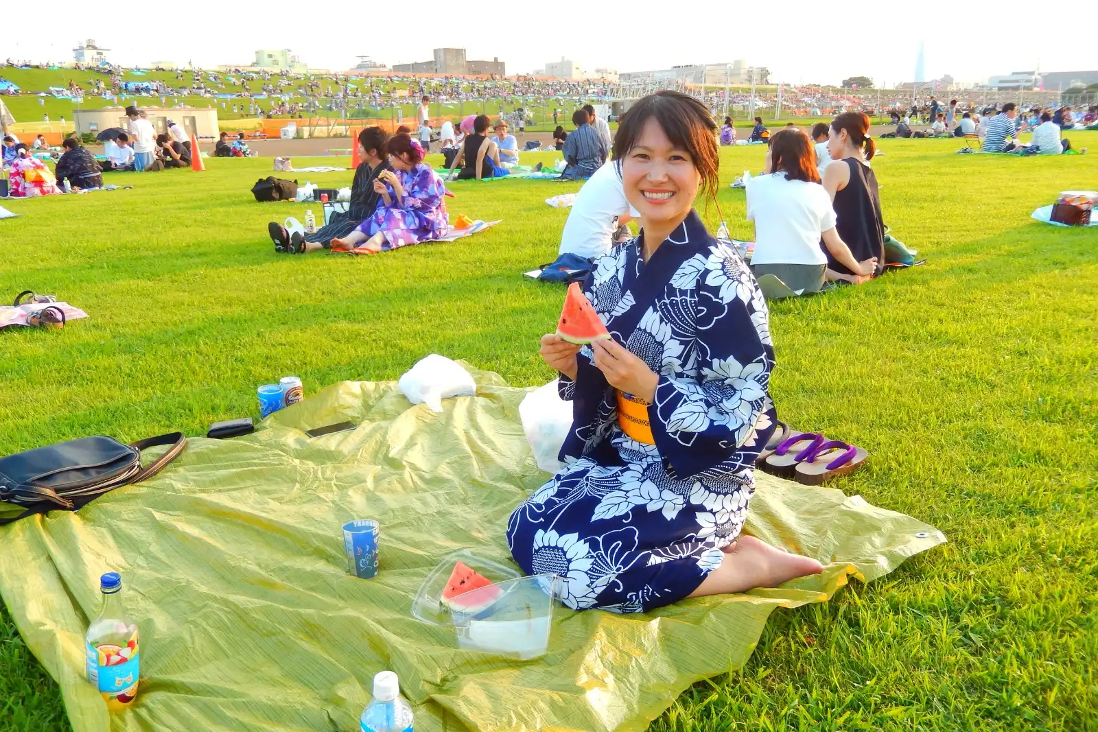 Hikari sits on a picnic sheet in a navy floral yukata, holding a slice of watermelon, with geta beside her and a crowded grassy slope behind her at an outdoor hanabi gathering.